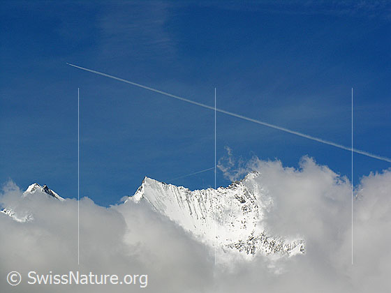 Foto: Durch Wolken sind Dom, Nadelhorn und Lenzspitze zu sehen. 
Darüber ein Kondensationsstreifen eines Flugzeugs.