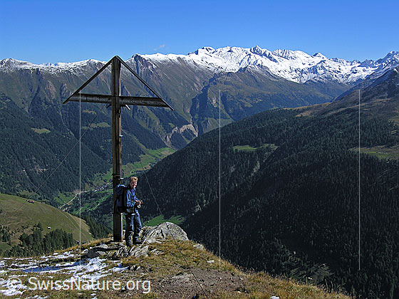 Foto: Wanderer bei Gipfelkreuz auf dem Meiggerhorn mit Aussicht über das Binntal und Tiefblick in den Talgrund. Die Gipfel (Rappehorn, Turbhorn, Hohsandhorn) sind bereits schneebedeckt.