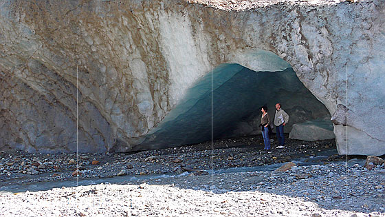 Foto: Zwei Personen bestaunen die Eismassen am Gletschertor des Steingletschers. Das Eis in der Wölbung ist türkis gefärbt und am Boden liegen grosse Eisstücke. Aus dem Tor fliesst nur wenig Wasser.