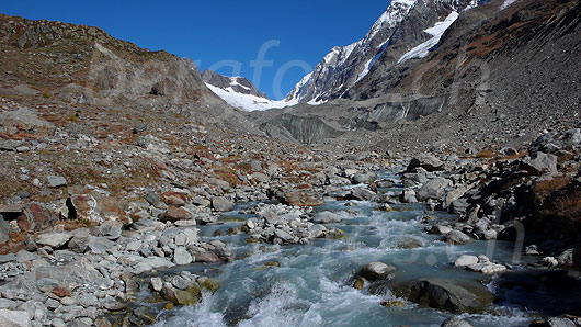 Foto: Lonza, Langgletscher und Lötschenlücke. Der Gletscherbach fliesst durch das Geröll durchsetzte und herbstlich gefärbte Gletschervorfeld des Langgletschers. Die zerklüfteten Eismassen des Gletschers sind mit Schutt bedeckt. Im Hintergrund sind Anuchnubel, Lötschenlücke und Sattelhorn zu sehen.