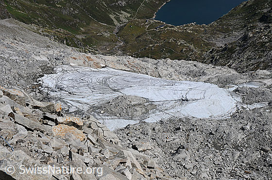 Foto: Lucendrogletscher (Ghiacciaio di Lucendro) umgeben von Felsblöcken und Geröll. Hintergrund: Tiefblick auf das Ufer des Lucendro Stausees.