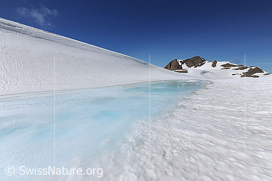 Foto: Schneebedeckte Hochebene mit zugefrorenem See und Berg im Hintergrund. Die Eisoberfläche ist angetaut und das Wasser über der Eisdecke erscheint hellblau.