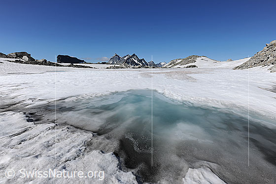 Foto: Weite Eisfläche eines zugefrorenen Bergsees mit Bergspitzen im Horizont. Im Vordergrund ist Wassereis des auftauenden Sees (Schwarzsee) zu sehen.