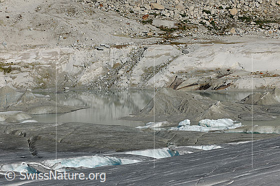 Foto: Eiskanal mit Eisbrücken im Gletschereis des Rhonegletschers. Angrenzend der Gletschersee mit Toteis, welches als Relief einer Gebirgslandschaft aus dem Wasser ragt. Im Hintergrund sind Sandablagerungen und Geröll zu sehen.
