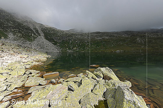 Foto: Wolkenstimmung mit Lichtspiel am Lengsee (oberhalb Obergesteln) bei vorwiegend bewölktem Himmel. Blockgelände bildet das Ufer des Bergsees. Die blaugrün schimmernde Wasserfläche befindet sich teilweise im Licht und teils im Schatten.