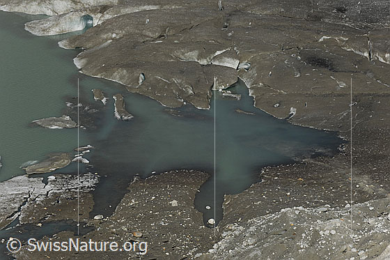 Foto: See im Gletschereis des Rhonegletschers. Der Gletschersee hat eine interessante Form angenommen und im Wasser befinden sich Eisschollen. Die Eisfläche ist mit Sand und Kies bedeckt.