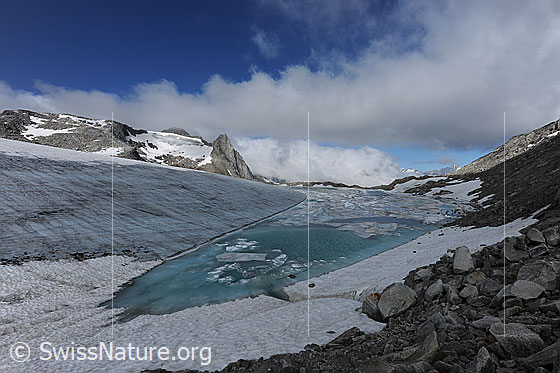 Foto: Gletscher und Gletschersee mit Wolkenstimmung. Urtümliche Gletscherlandschaft bestehend aus Blockgelände, Gletschersee im Gletschereis und Eisschollen, welche sich von der aufgebrochenen Eisdecke gelöst haben. Das Wasser hat die Farbe türkis. Durch das Abschmelzen des Chüebodengletschers vergrössert sich der See laufend (Gletscherschwund, Klimawandel). Mittlerweile misst der See in der Breit ca. 100m. Im Jahr 2008 waren erst zwei sehr kleine Seen vorhanden.
