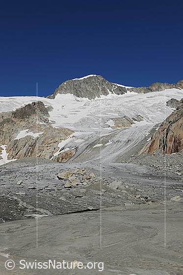 Foto: Gletschervorfeld Tiefengletscher und Galenstock. Darüber wolkenlos blauer Himmel. Im sandigen Gletschervorfeld sind Wasserläufe zu sehen. Die Gletscherzunge endet flach und führt Geröll und Felsblöcke mit.