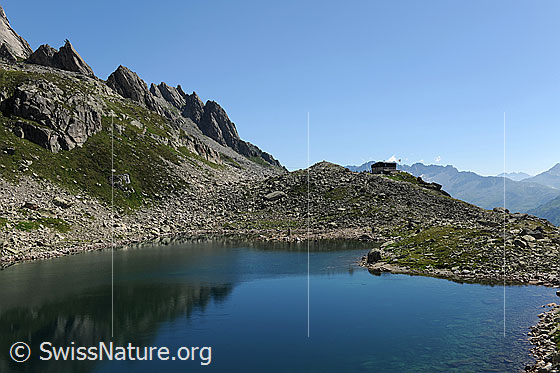 Foto: Bergsee und Bergseehütte. In der blauen Wasserfläche spiegeln sich Felszacken.