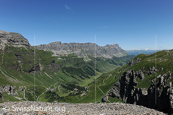 Foto: Urnerboden, Läckistock, Jegerstock und Ortstock.
Tiefblick auf die Passstrasse zum Klausenpass und auf den Urnerboden im Taleinschnitt. Die Berghänge und Alpweiden sind grün. Im Hintergrund ist das Felsmassiv mit Läckistock, Jegerstöck und Ortstock zu sehen.