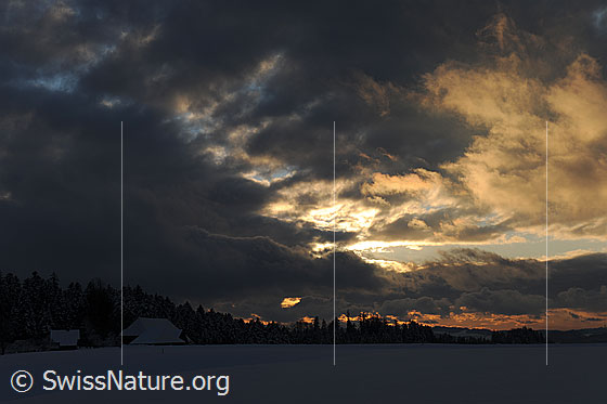 Foto: Wetterfront mit Abendrot über Winterlandschaft im Emmental. In der Dämmerung ist eine weite, offene Schneefläche mit Bauernhof und verschneitem Wald zu sehen.