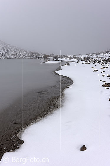 Foto: Kleiner Bergsee mit schneebedecktem Ufer im Nebel.